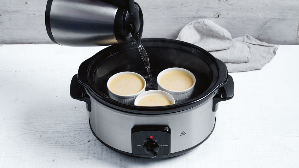 Kettle pouring water into a slow cooker holding 3 baked custards in small ceramic bowls